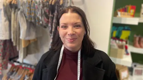 Emma Moore standing in her shop. She has dark brown hair and is wearing a black jacket. She has a measuring tape hung around her neck and there are clothes hanging up behind her.