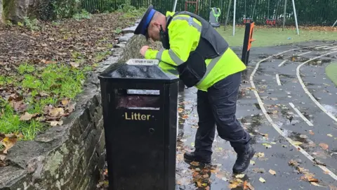 A PCSO peers into a litter bin in a park with swings in the background. There are leaves on the ground. 