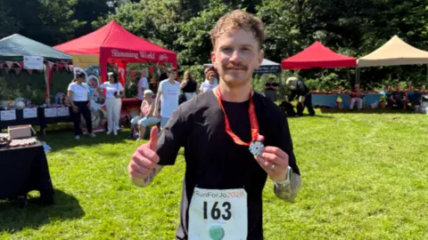 Jonathan Reed/BBC A smiling man doing a thumbs up gesture and holding a medal on a red ribbon around his neck. He is wearing a black t-shirt with a race label on which says 'Run For Jo 163'.