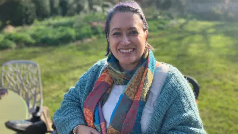 ME patient Radha smiling for the camera as she sits on a chair outside. She is dressed in woolen clothes and wrapped up with a colourful scarf.