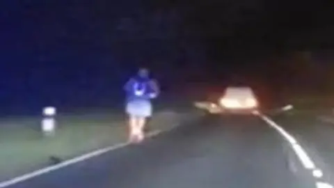 The e-scooter rider travelling on the A-road wearing a purple T-shirt and grey shorts. He is on the left of the road at night time and it is dark. A car is being driven in front of him. The picture appears to be a still taken from dashcam footage.