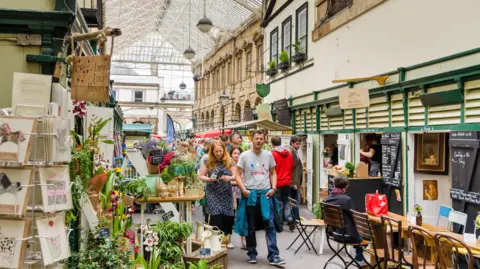 Getty Images St Nicholas Market in Bristol. Customers can be seen walking through the indoor market, looking at vendors. Greeting cards and plants can be seen on the left hand side of the frame, while tables and a food stall can be seen on the right. 