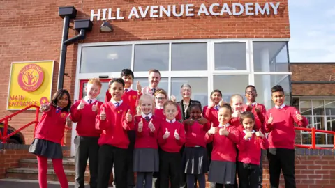 Fourteen pupils, boys and girls, are wearing red school jumpers and are smiling with their thumbs up. Behind them are two teachers. They are all standing in front of the red brick of a school building. There is a large silver sign on the wall reading "Hill Avenue Academy"