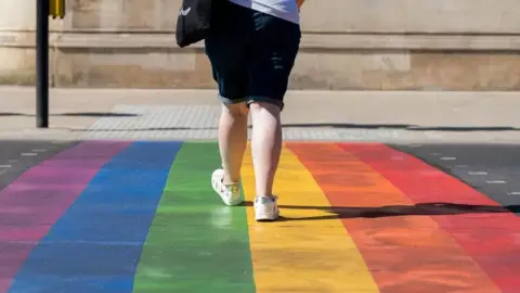 Person wearing denim shorts walking across rainbow pride flag crosswalk