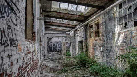 A derelict building with windows smashed up and overgrown weeds. There is also graffiti on the walls 