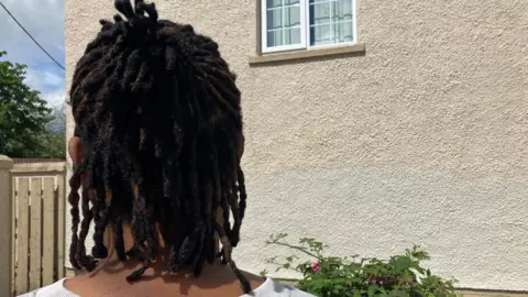 Uche Ukeje stands facing the gable wall of her house and with her back to camera. She has black hair and is wearing a white jumper. A window of the house is visible as is a garden plant, and fence.