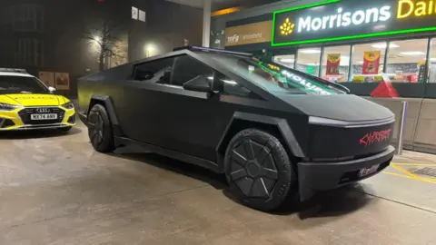 GMP The black Tesla Cybertruck parked up at a petrol station next to a Morrisons Daily branch and in front of a yellow liveried police car