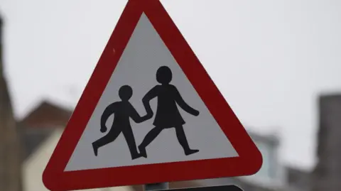 BBC A school sign, crossing patrol warning. The warning sign is in a triangular shape. It has a red border and a white background with two figures portraying a woman and child walking while holding hands.