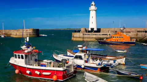 The image shows Donaghadee harbour with a white lighthouse in the background. In the foreground are a number of colourful boats.