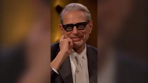Jeff Goldblum smiles. He is wearing a white shirt and tie with a dark grey jacket, and black glasses with thick rims.
