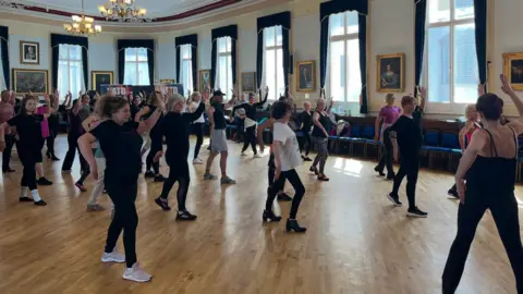 A lively group session taking place in a spacious, traditional hall with wooden floors and chandeliers. Participants are arranged across the room, moving in coordinated ways as part of a guided physical activity. The setting is bright, with large windows and framed artwork lining the walls, creating a welcoming and energetic atmosphere. Attendees of varied ages are dressed in comfortable clothing suitable for dancing. 