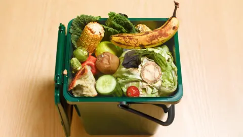 A green kitchen waste bin with its lid opened, loaded with a banana, sweetcorn, lettuce and a cucumber, placed on a wooden kitchen floor.