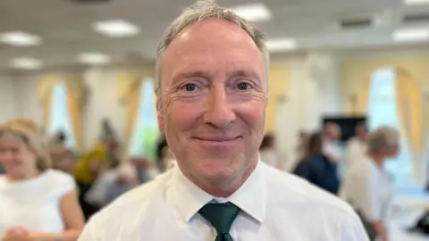 Paul Montague looks at the camera. He has grey hair and a white shirt with a green tie. 