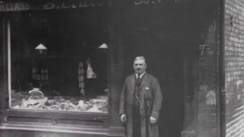Manchester Jewish Museum A man in an overcoat, shirt and tie stands in the doorway outside of a butchers.