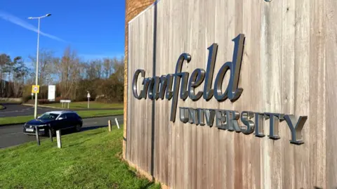 Martin Heath/BBC A road junction with a large sign showing the words Cranfield University in silver lettering with black borders on a background of wooden slats. There is grass in front of the sign, and a dark-coloured car is heading past the sign on the adjacent road. There is grass bordered by trees on the opposite side of the road.