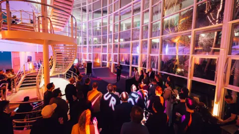 Kinrise / ShotAway A crowd gathered in a large room with a high ceiling and metallic staircase. The walls are covered in glass panelled windows and it is dark outside. The indoors is lit up with pink, purple and orange hues. The crowd of people are all facing towards a short stage, where a man is giving a speech.