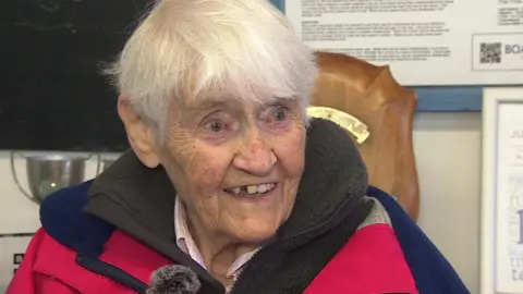 A smiling Jean Paton. She has short white hair and is wearing a red and blue sailing coat.