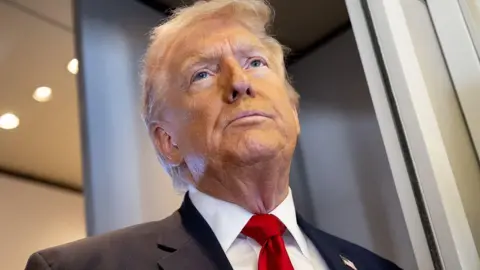 Getty Images Donald Trump speaks with the media aboard Air Force One during a flight. He is wearing a black suit and red tie