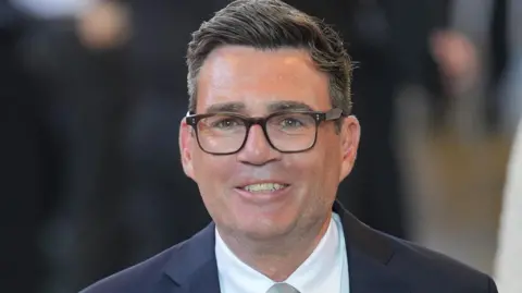 Andy Burnham smiling and looking towards the camera. He is wearing dark rimmed glasses, a dark suit jacket, white shirt and tie. He has dark hair, swept to one side. He is prominent in the centre of the image and the background is blurred.