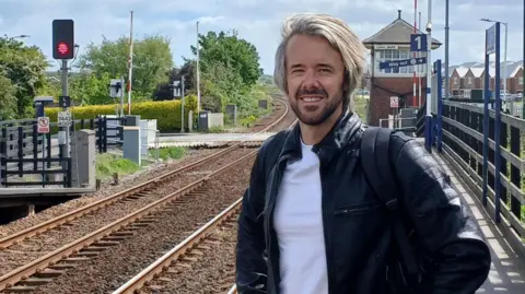 Tristan Learoyd, a man with a dark stubble beard and a mane of blonde hair, standing next to the railway line at Marske train station. He is wearing a dark coat and white t-shirt. Behind him, the line curves past a road crossing and into the greenery beyond.