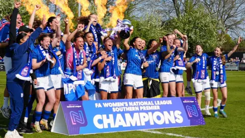Ipswich Town Women celebrate in a group on a football pitch. A banner sits in front of the team that reads: "FA Women's National League Southern Premier Champions." Flames can be seen shooting in the air behind them.