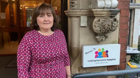 A woman looking into the camera. She is wearing a pink, red and black geometric patterned dress. She is standing next to a sign that reads: "Welcome to Nottinghamshire Hospice. Adding life to days".