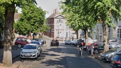 Google A street with cars and trees along both sides and a tall house in the background