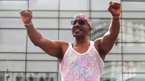 Mr Motivator standing in front of a glass building. He has both of his arms in the air and is wearing a white string vest over a brightly coloured tank top. He is smiling and wearing a bandanna and sunglasses.