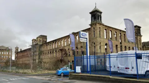 A light-brown coloured shell of a former mill building with scaffolding on two towers at the front of the structure and black charring on another tower's turret evidence of fire damage. 