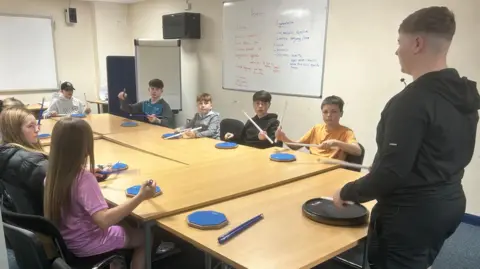 Eight children, sat around a table with drum plates and sticks, an older male is stood at the end of the table, demonstrating the instrument