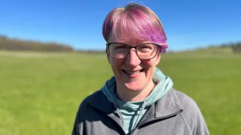 A woman with pink highlighted hair and dark glasses stands in a field wearing a grey hoodie and fleece.