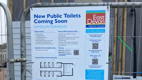 A sign saying "new public toilets coming soon" at Exmouth Foxholes, posted on wire fencing in front of the construction site 