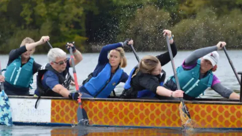Five men and woman paddle in a dragon boat on a calm expanse of water. The boat is painted orange and yellow and the paddlers are wearing blue life jackets and are paddling either side of one another.