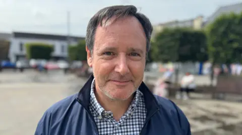 BBC A man with dark grey hair and blue eyes, white and blue checkered shirt and navy lightweight jacket smiles at the camera with a blurry image behind him of cars and buildings.  