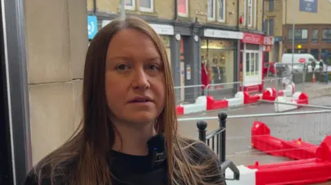 Nathan Turvey/BBC A picture of Kirsty Devine who has brown hair standing in her shop doorway with roadworks in the background.