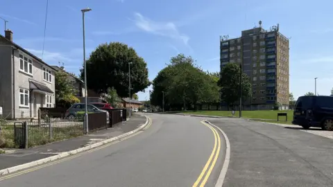 A quiet residential street with a grey detached house, clean pavements, some trees, and a tower block in the distance.
