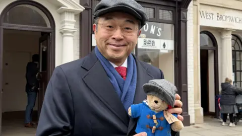Hiroshi Suzuki has a grey flat cap on and a navy blue coat, with a blue scarf and red tie. He wears glasses and is holding a small Paddington Bear toy which also has a grey flat cap on, and a blue coat.