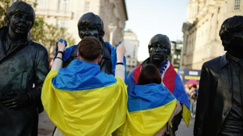 Two people clad in Ukrainian flags drape flags around the Beatles statues