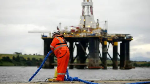 Matthew Lloyd / Bloomberg via Getty Images A person wearing orange reflective work gear and a white construction helmet pulls a blue rope while standing on the edge of the water as a mobile offshore drilling unit is transported into the harbour in the Port of Cromarty Firth in Cromarty in 2016.