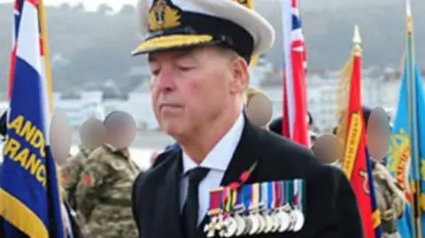 man dressed as a navy officer with medals pinned to his chest and surrounded by other military personnel and colourful flags