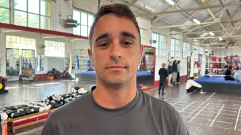 Joe is wearing a grey top with Release Jersey written on it in white. He has short, brown hair. He is stood in gym with a blue boxing ring behind him on the right.