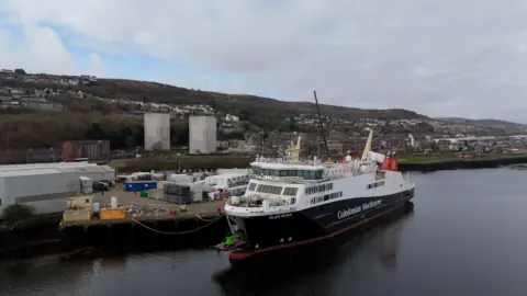 A Caledonian MacBrayne ferry moored at a dock in Inverclyde, with industrial buildings and hillside housing visible along the waterfront.