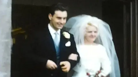 BBC A grainy wedding photo - on the right Beryl, who has blonde hair, is wearing a white wedding dress and a white veil. On the left, Griff has dark hair and is wearing a dark suit, with a light blue tie and waist coat. 