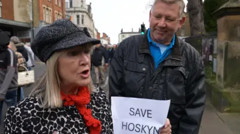 Michelle Mitchell, with blonde hair, is wearing a black and white hat and coat and is carrying a Save Hoskyn piece of paper. She is standing next to a man and people are marching in the background.