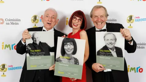 IMROs Hugo Duncan at the IMRO awards wearing a suit and holding his Hall of Fame Plaque, he is standing beside Carol Dooley (dressed in red with red glasses and dress) and Eddie Caffrey in a black suit.