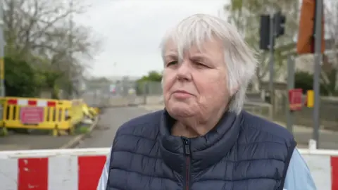 Dawn Johnston wearing a body warmer while stood outdoors on a road. The road appears blocked and there is temporary traffic control equipment visible behind her.