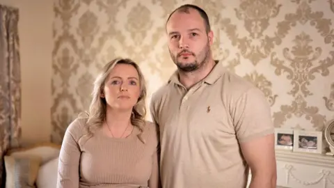 Heidi and Dale standing close together in a living room with cream patterned wallpaper behind them. Pregnancy scan photos are framed on the fireplace. Heidi has long, wavy blonde hair and is wearing a ribbed, long sleeved top and gold necklace. Dale has short dark hair with a beard and is wearing a cream polo-shirt.