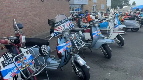 a row of scooters, without their riders, are lined up in a car park