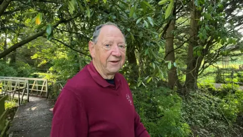 Handout Mr Nettle stood in a red top near a bridge and green bushes. He has blue eyes and white hair.