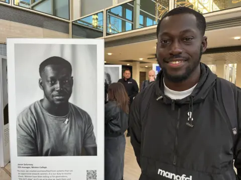Nicola Haseler/BBC Jason Selormey, a young man with short hair and a beard, smiles as he stands next to his portrait, which is featured in the exhibition. Jason is wearing a black top with a white ribbon pinned to it. He is also wearing a white ribbon in the portrait. The gallery showing the exhibition is in the background and people can be seen looking at the pictures.